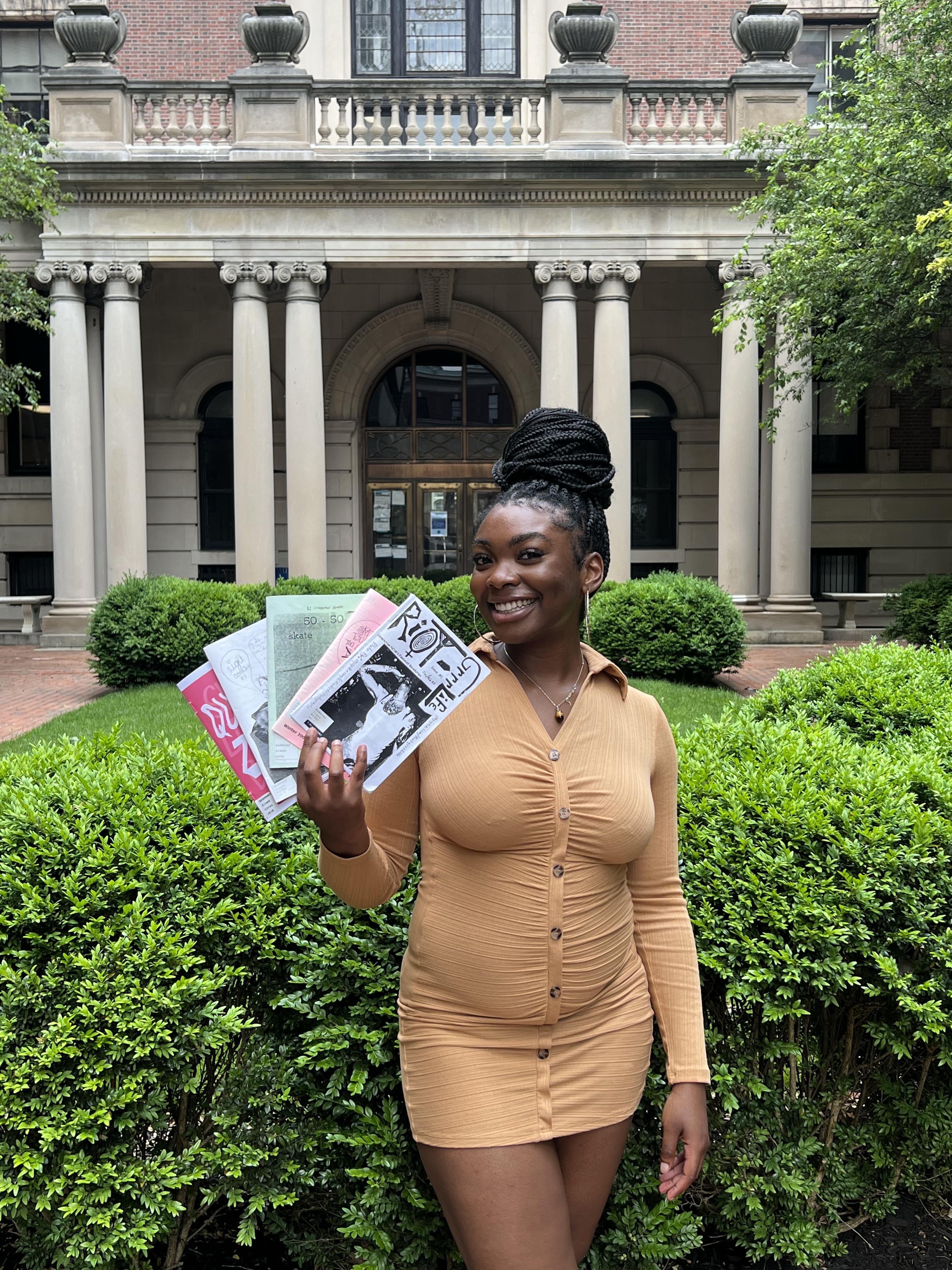 the curator of the exhibit, Erinma, in tan long sleeve dress holding colorful zines in her hands